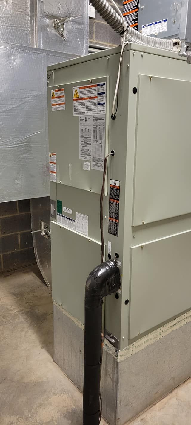 Gray HVAC unit in a basement mechanical room with black pipes and ductwork.