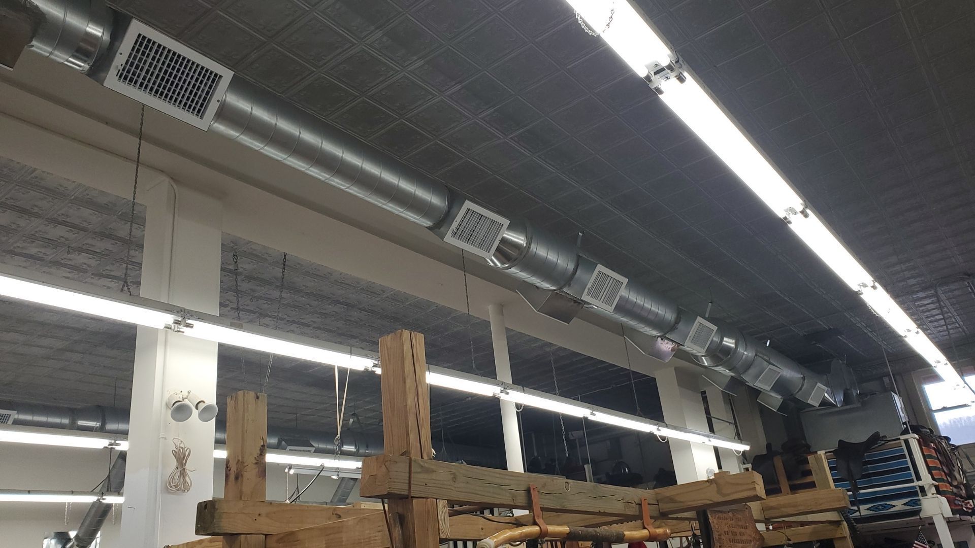Warehouse aisle with stacked wooden pallets, metal shelving, and overhead ventilation ducts under bright lights