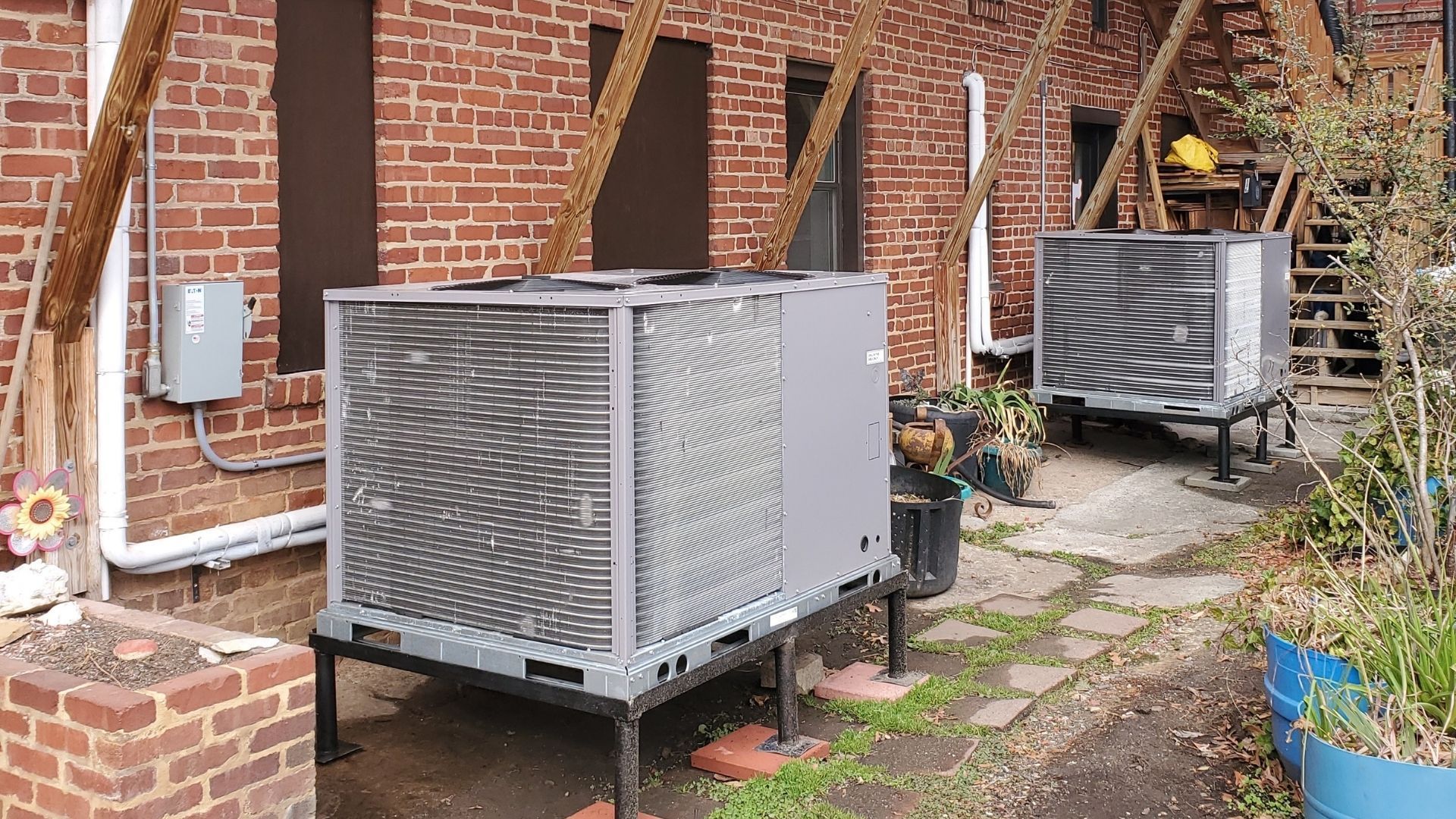 Outdoor HVAC units beside a brick house, with one unit in the foreground and another along a stone path.