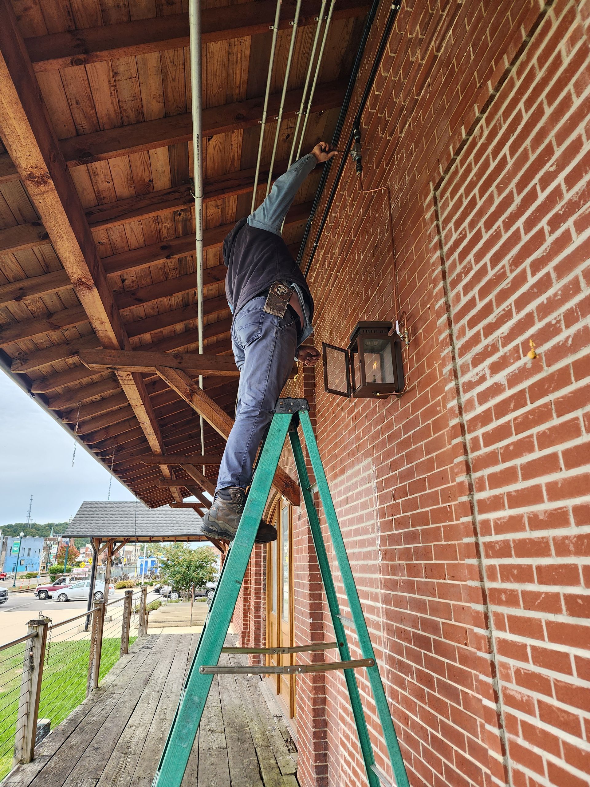 Person on a green ladder adjusting overhead cables beside a brick wall under a wooden roof