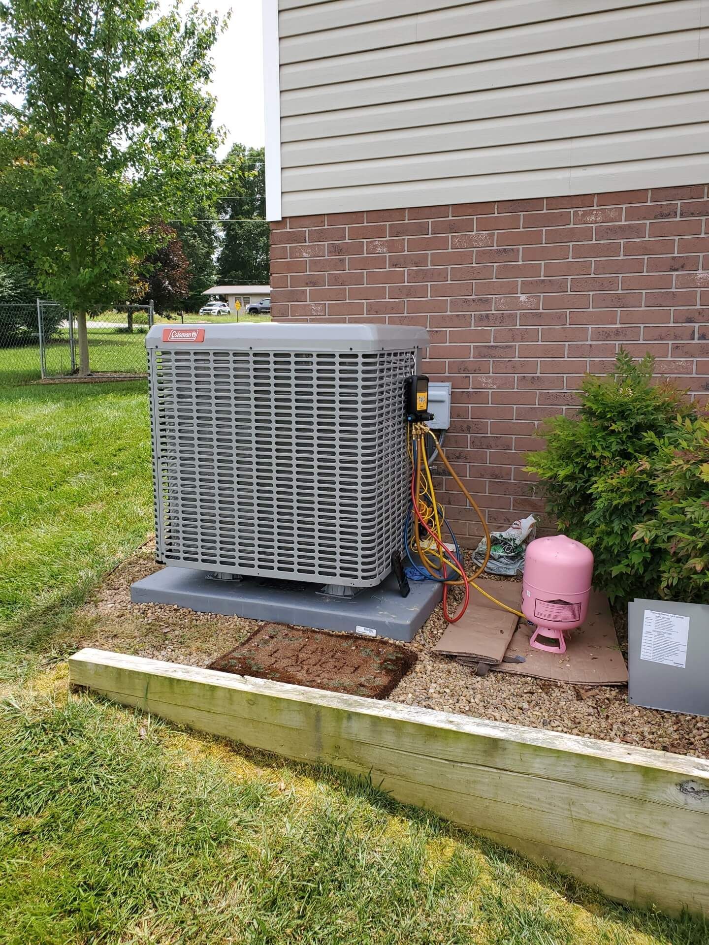 Outdoor HVAC unit beside a house, with a pink propane tank and small shrubs in a yard.