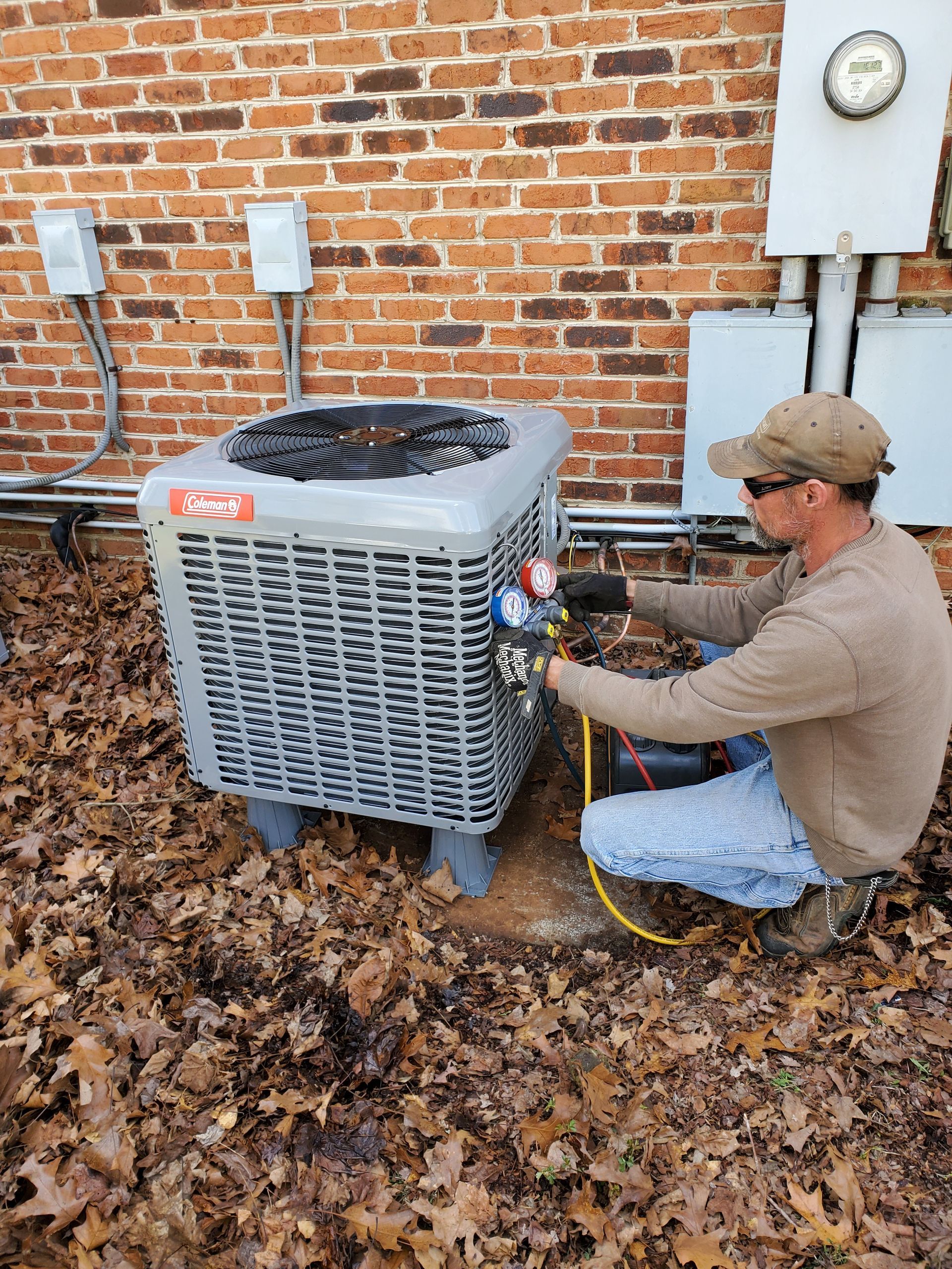 Contact Us Technician kneeling beside outdoor HVAC unit, checking gauges against a brick wall in a leaf-covered yard.