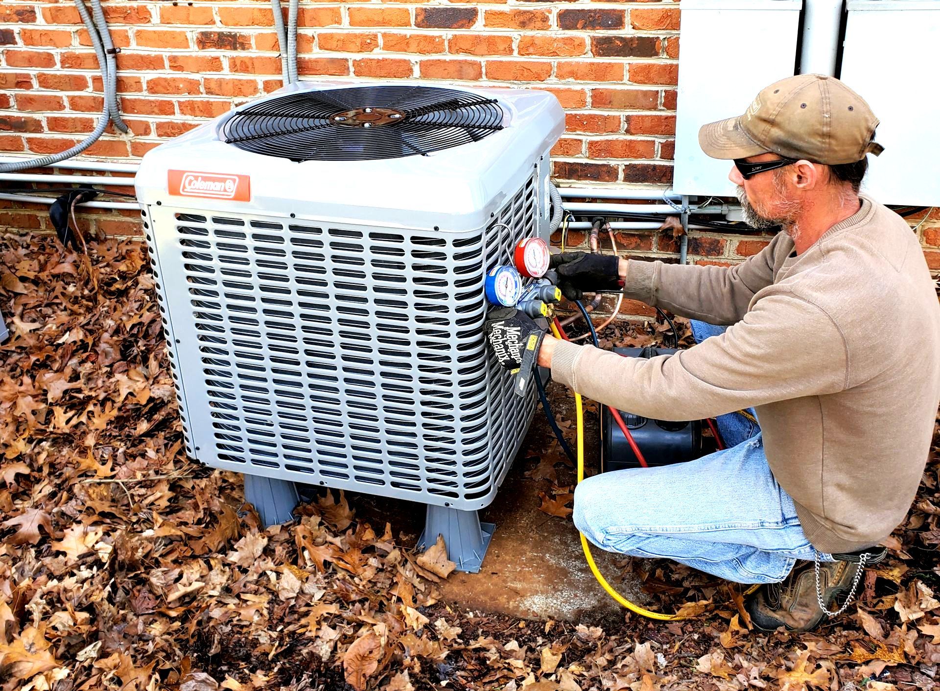 HVAC Services Technician servicing an outdoor air conditioner unit beside a brick wall in a yard covered with leaves