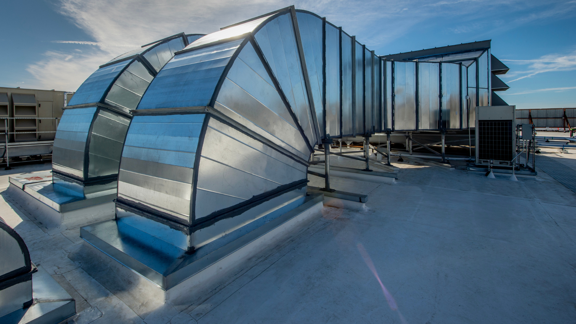 Curved metal rooftop ventilation ducts and glass skylight structures on a flat roof under blue sky