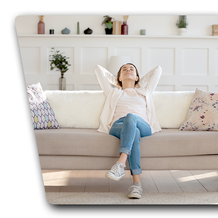 Contact Us Woman relaxing on a beige sofa in a bright living room, eyes closed with arms behind her head.