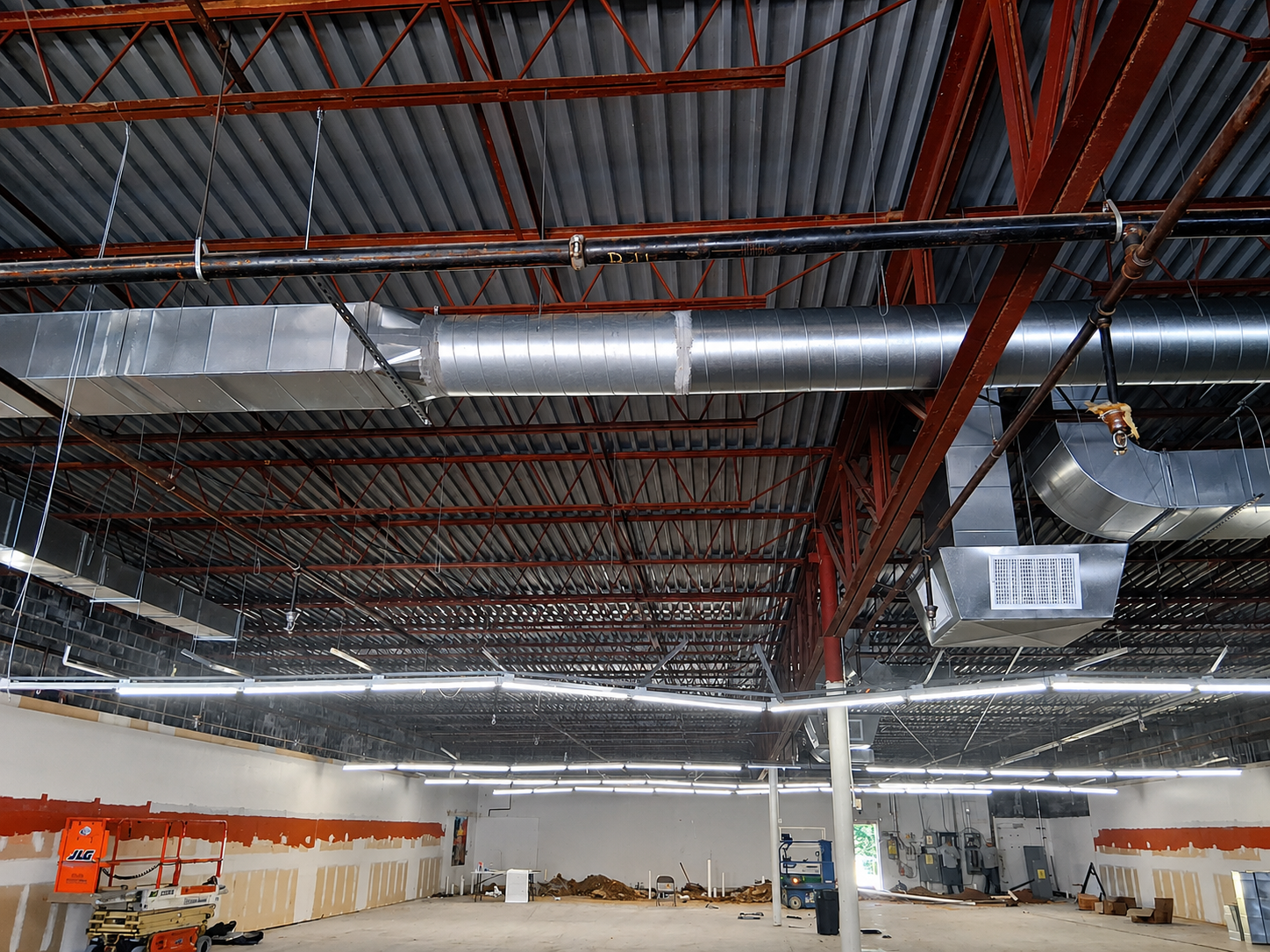 Warehouse ceiling with exposed red beams, silver ductwork, and bright overhead lights above an open floor.