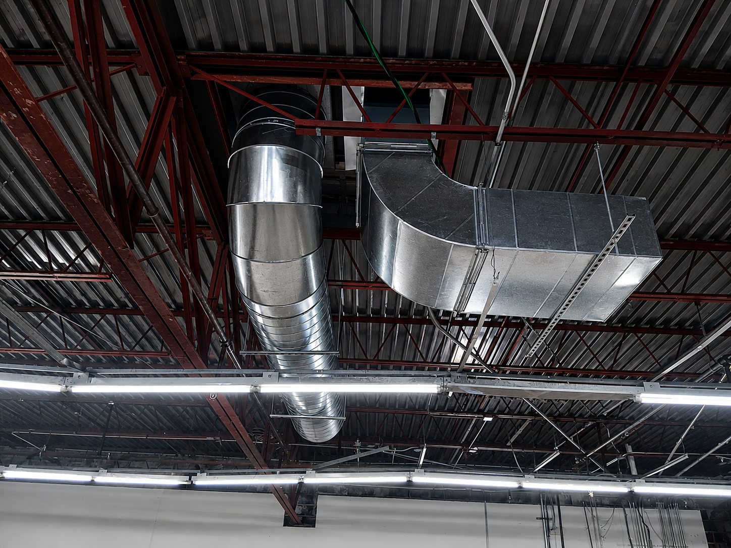 Industrial ceiling with silver HVAC ducts, red pipes, and fluorescent lights in a warehouse interior