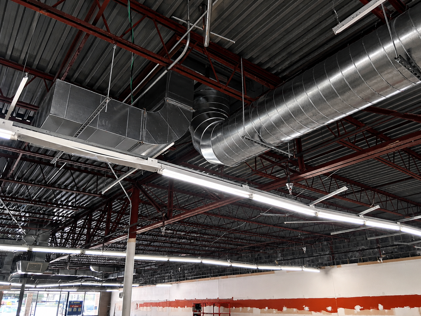 Ceiling ductwork and red steel beams in an unfinished industrial interior with white walls and exposed wiring