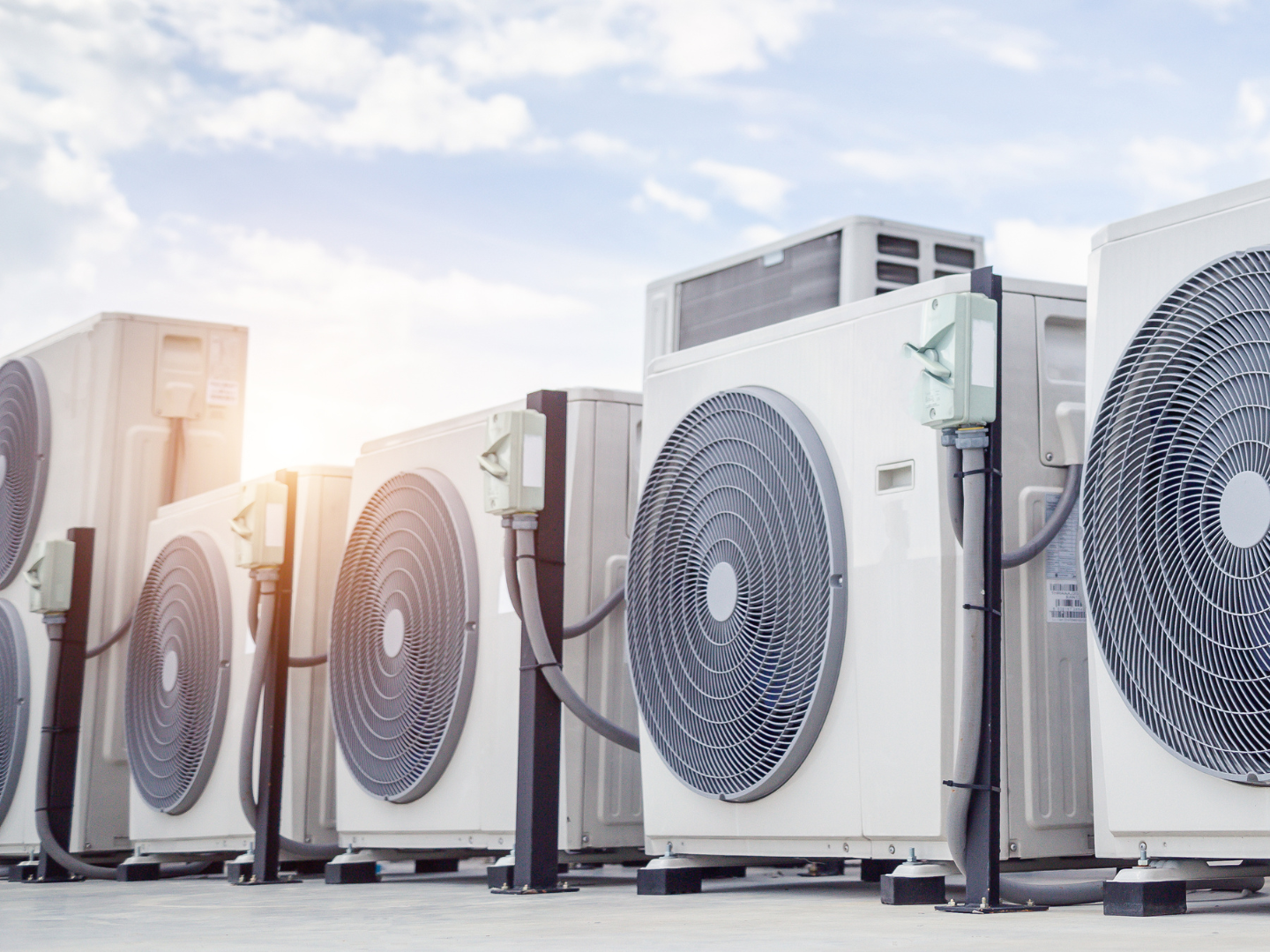 Row of outdoor air conditioning units with large fans beside a building under a bright sky