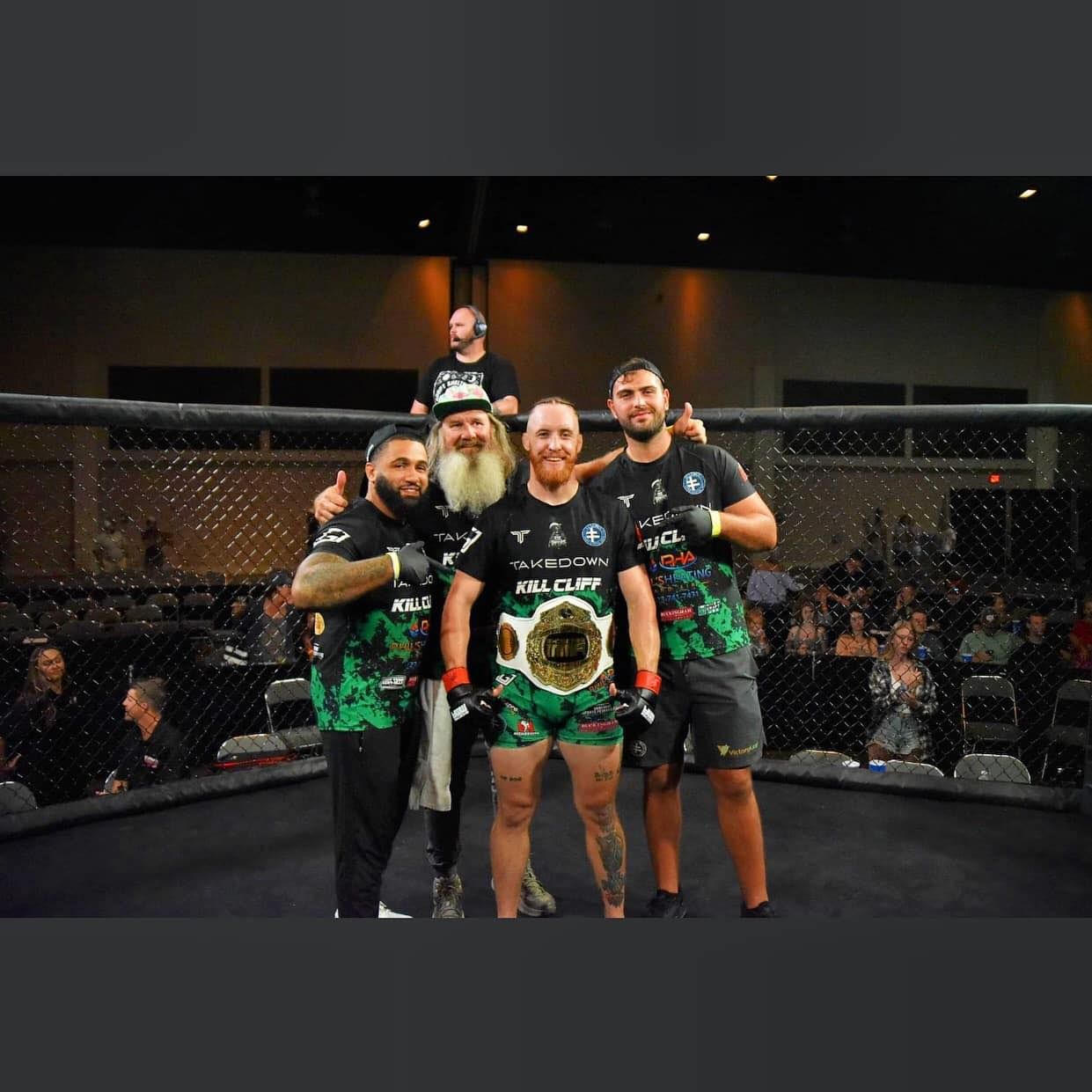 Four MMA fighters pose in a cage, one holding a championship belt, with a referee behind them.