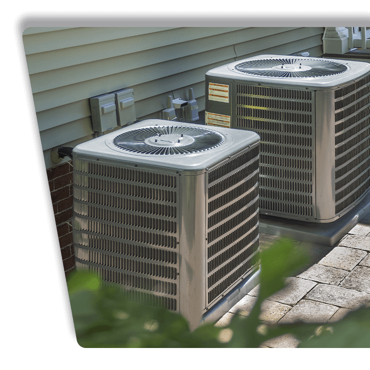 Two outdoor air conditioner units beside a house wall on a paved patio.