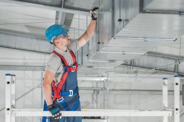 Contact Us Worker in blue helmet and red harness installing ceiling equipment in an industrial building