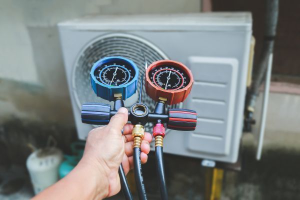 Hand holding HVAC manifold gauges with blue and red dials in front of an outdoor air conditioner unit
