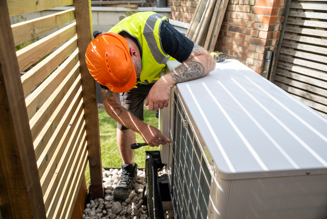 Worker in orange hard hat inspecting an HVAC unit beside a wooden fence and house siding.