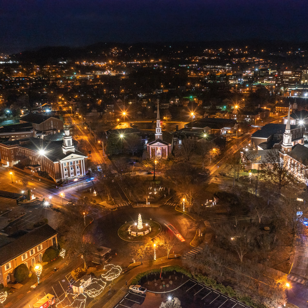 Nighttime aerial view of a brightly lit town square with buildings, trees, and city lights