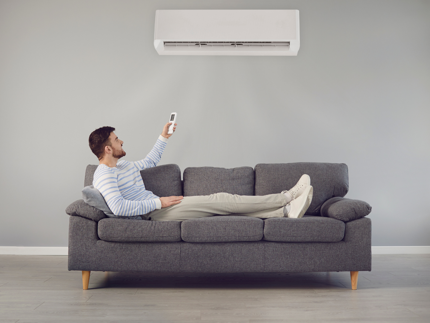 Man relaxing on a gray sofa, pointing a remote at a wall-mounted air conditioner in a minimalist room