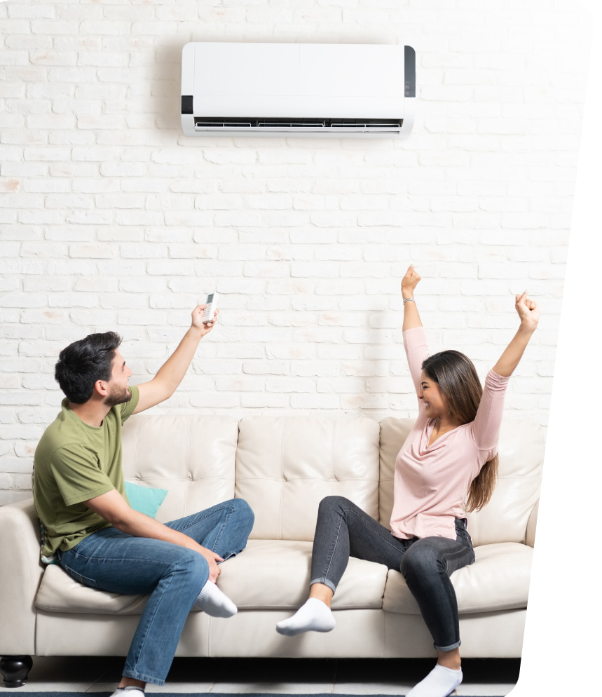 Two people on a couch using a remote to adjust a wall-mounted air conditioner in a bright living room.