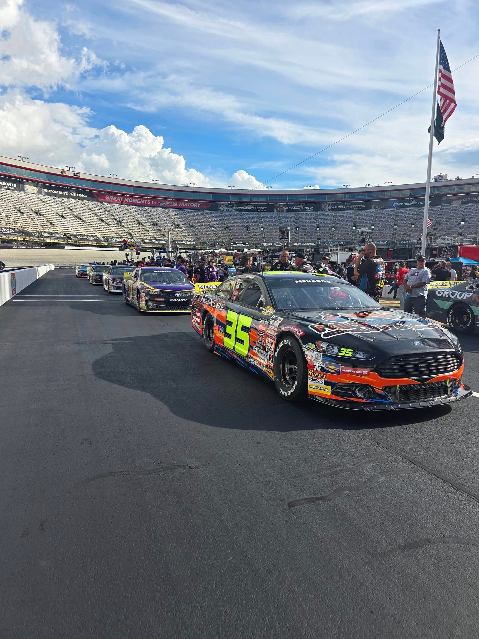 NASCAR race cars lined up on the track in a stadium, with an American flag waving overhead.