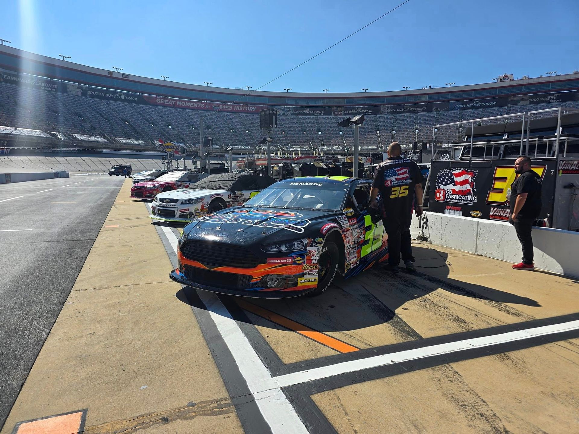 NASCAR pit lane with black-and-orange race car and crew beside it at a stadium track