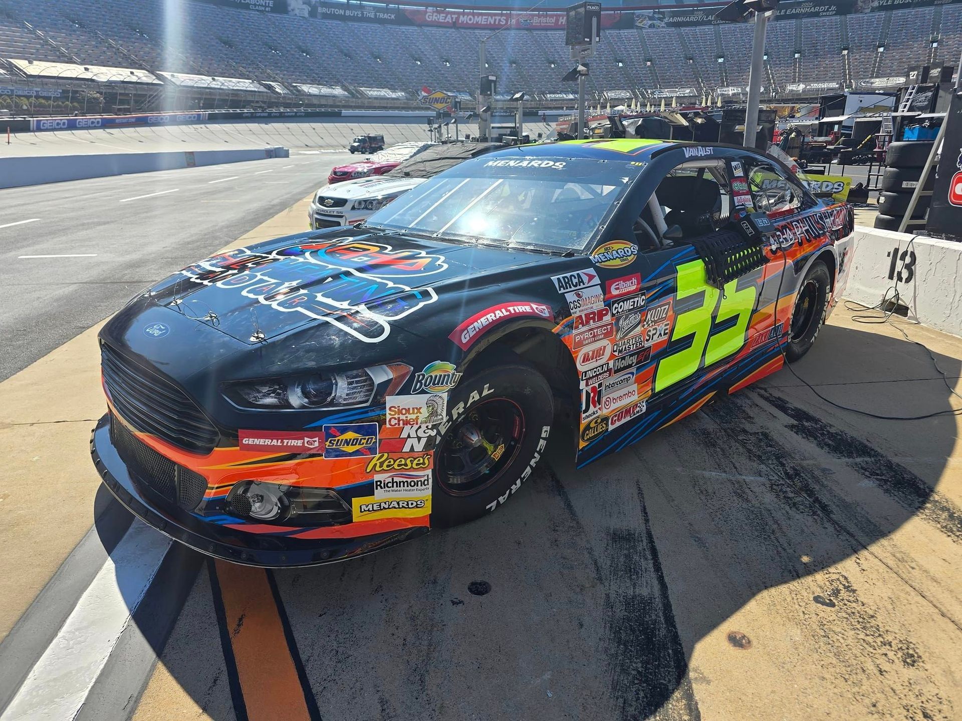 Black and neon green stock car in pit lane at a sunny racetrack, front view with sponsor decals