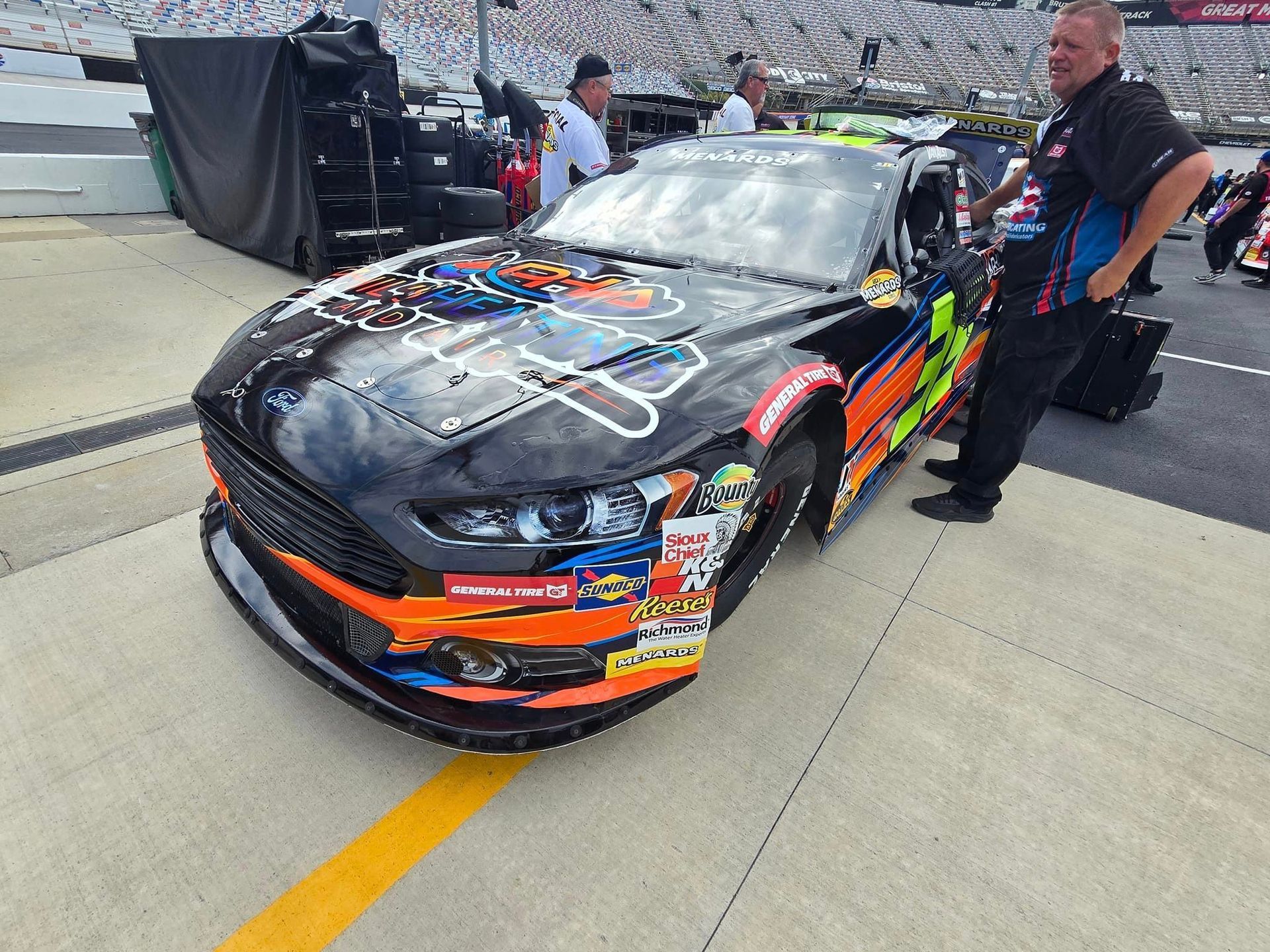 Damaged black race car on pit lane with crew members nearby and grandstands in the background