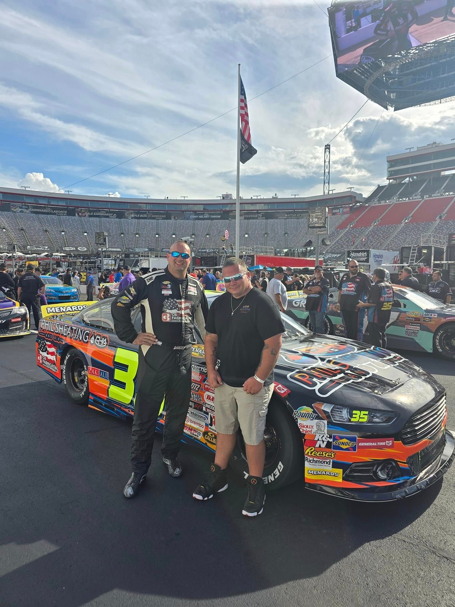 Two men pose beside a black race car on a track, with a stadium crowd in the background.
