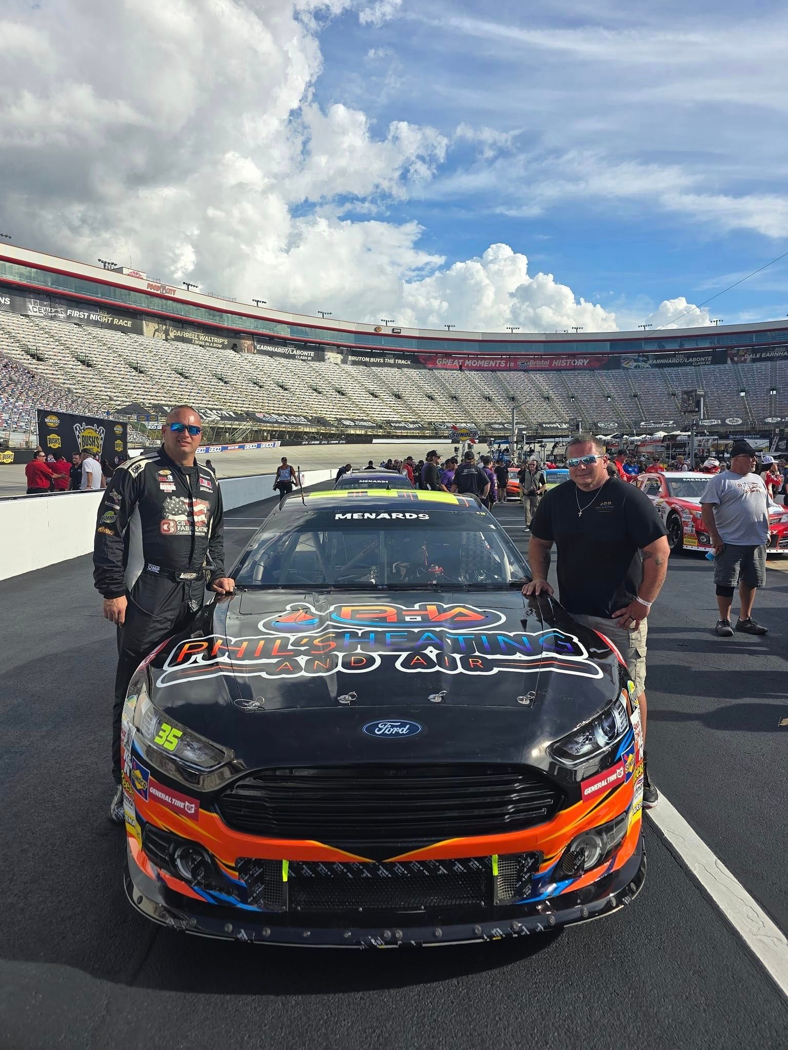 Race car on the track with two men standing beside it at a stadium under cloudy skies