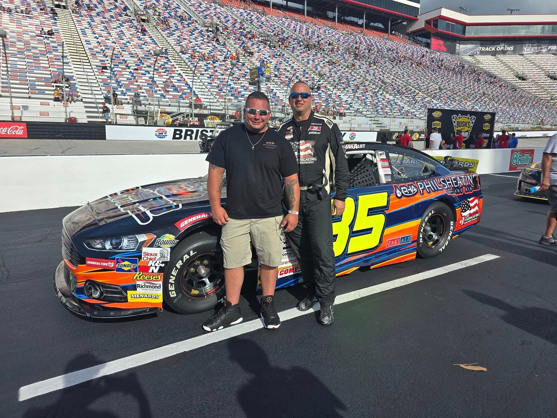 Two men posing beside a black-and-orange race car with number 35 on a racetrack, grandstands in the background