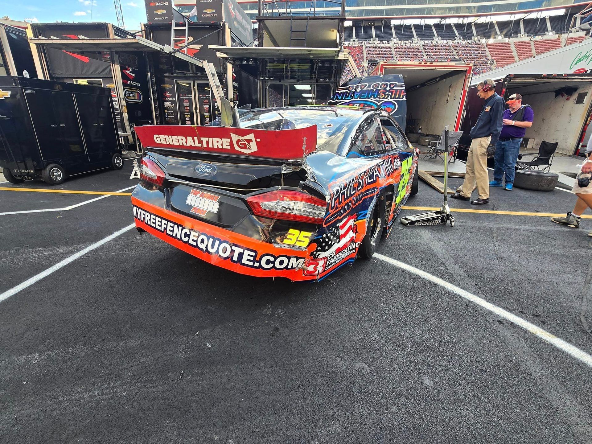 Damaged race car in a pit lane, front bumper crushed, with crew members beside it in a stadium.