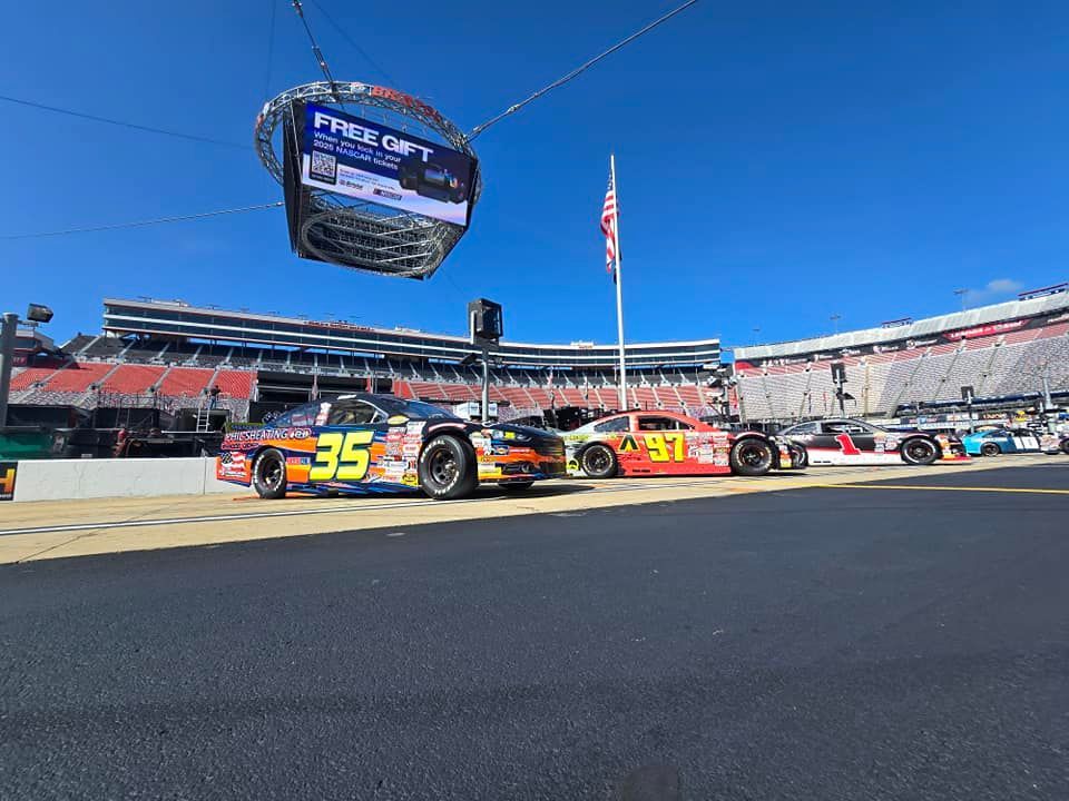 Two NASCAR stock cars on pit road at a racetrack with a hanging scoreboard and American flag