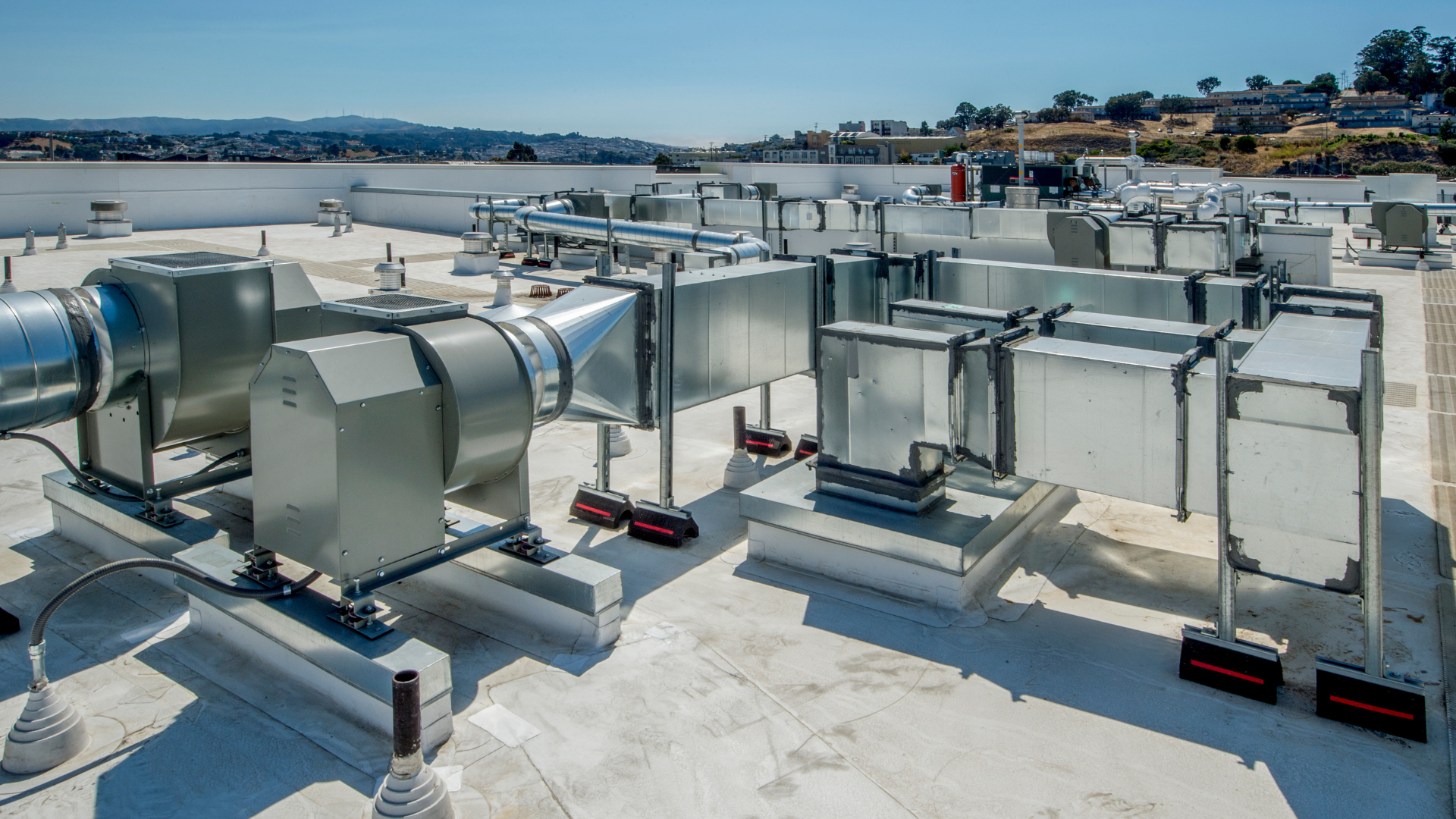 HVAC equipment on a rooftop, featuring silver ducts and machinery against a blue sky, cityscape in the background.