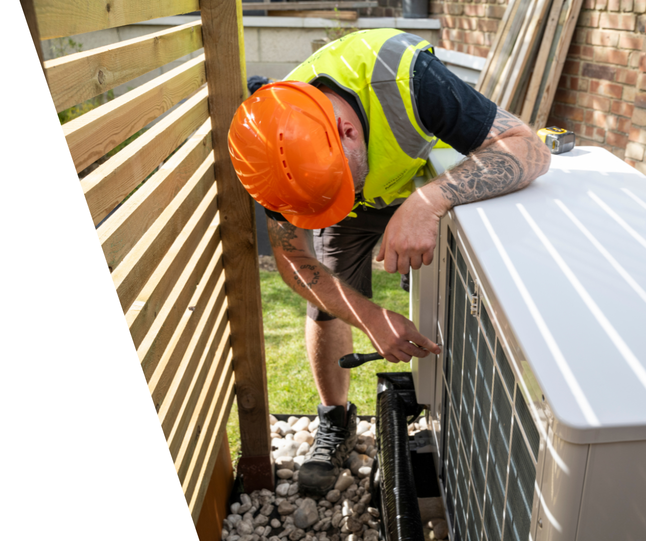 Contact Us! Worker in orange hard hat inspecting outdoor HVAC unit beside a wooden fence.