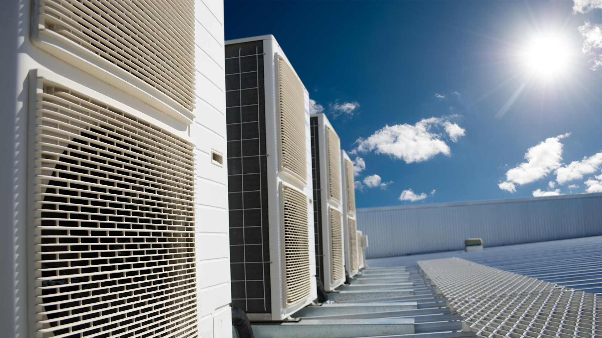 Row of rooftop HVAC units beside a bright blue sky and sunlit metal walkway