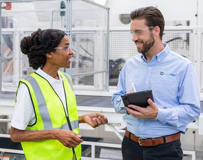 Two workers in a factory discussing with a tablet and clipboard, one wearing a yellow safety vest.