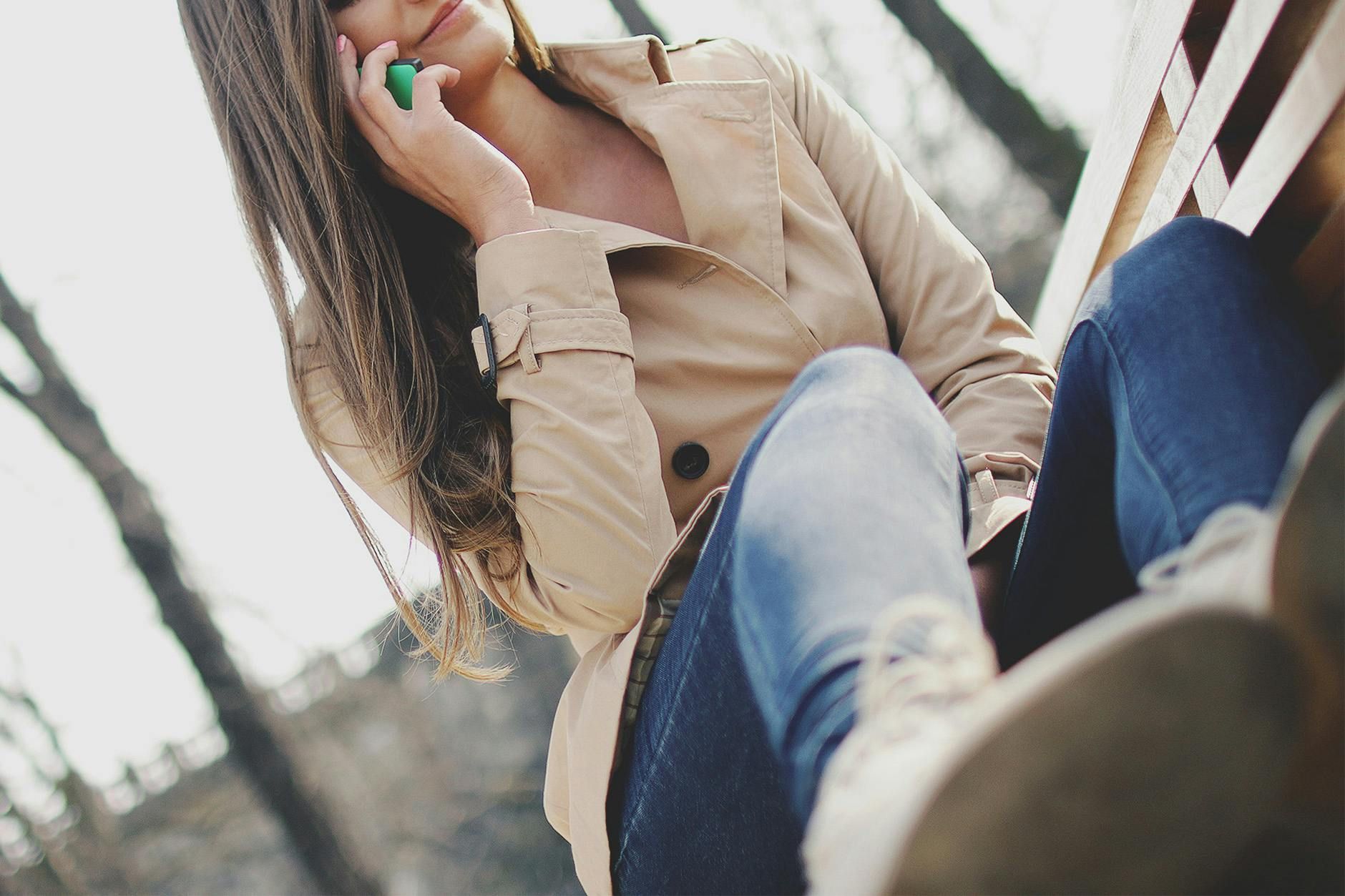 Woman sitting outdoors, talking on a cellphone, wearing a beige jacket and jeans