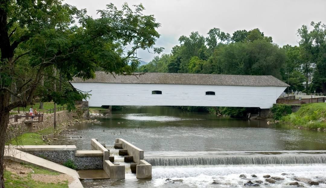 Covered watermill beside a small river dam, surrounded by green trees and cloudy sky