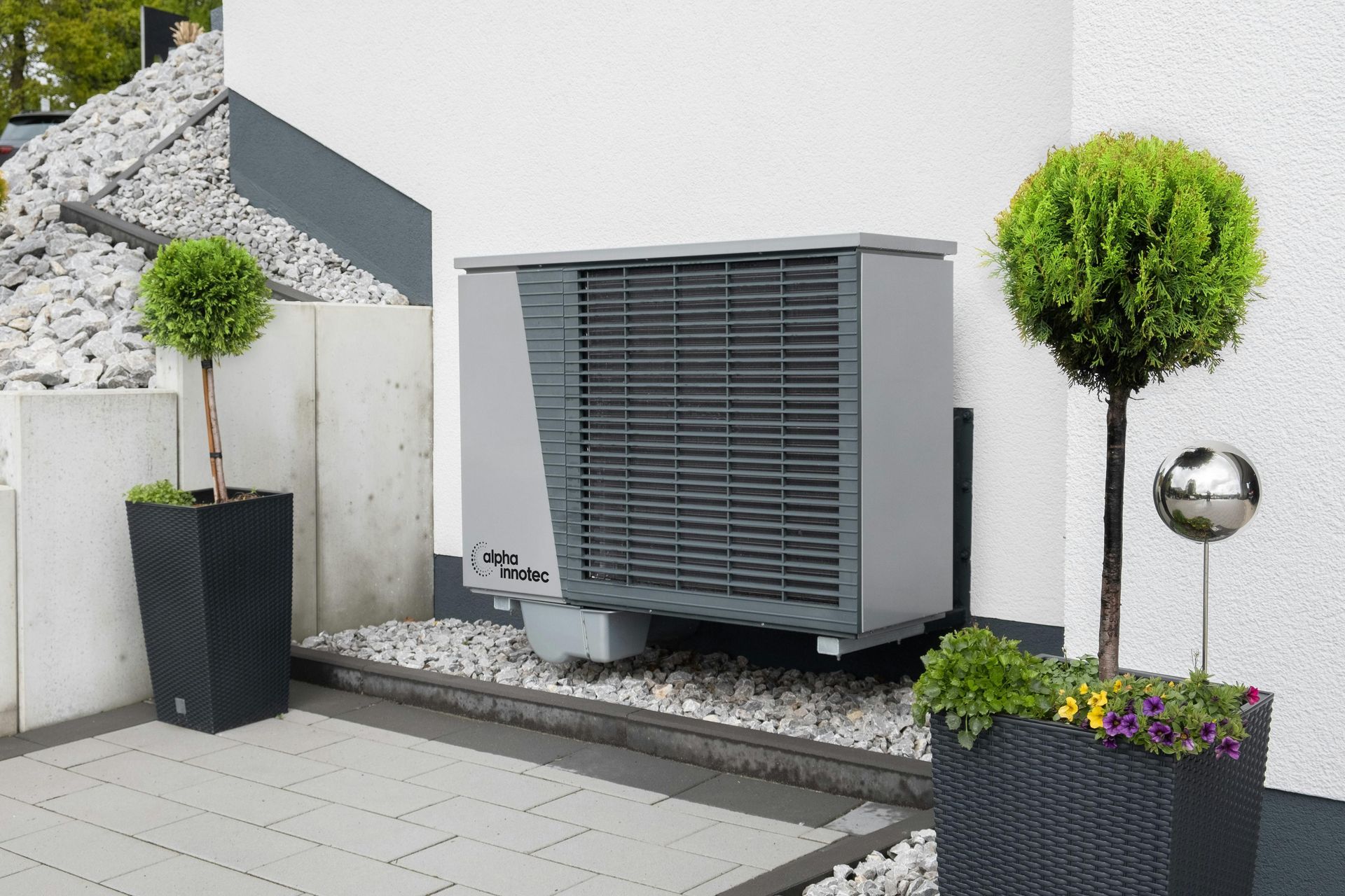Heat pump on a white wall with two potted trees and decorative stones.
