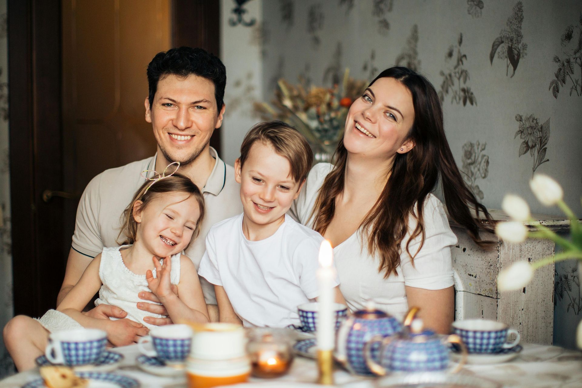 A happy family of four sitting around a table set for tea with white flowers in the foreground.