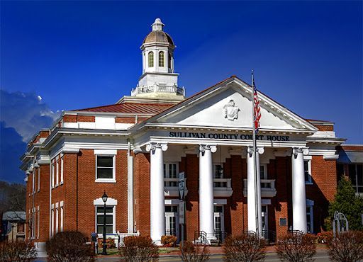 Red-brick civic building with white columns, a cupola, and a flag flying in front.