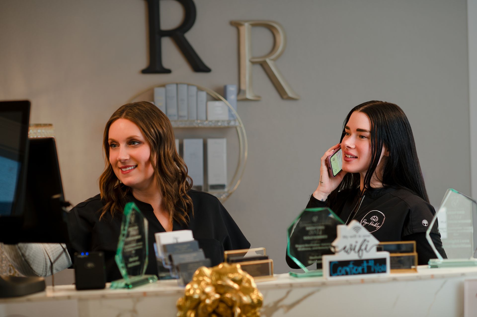 Two women are standing at a reception desk talking on their cell phones.