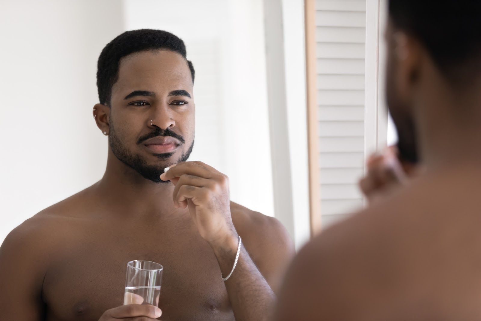 Man applying shaving cream to his face in a bathroom mirror, holding a glass cup