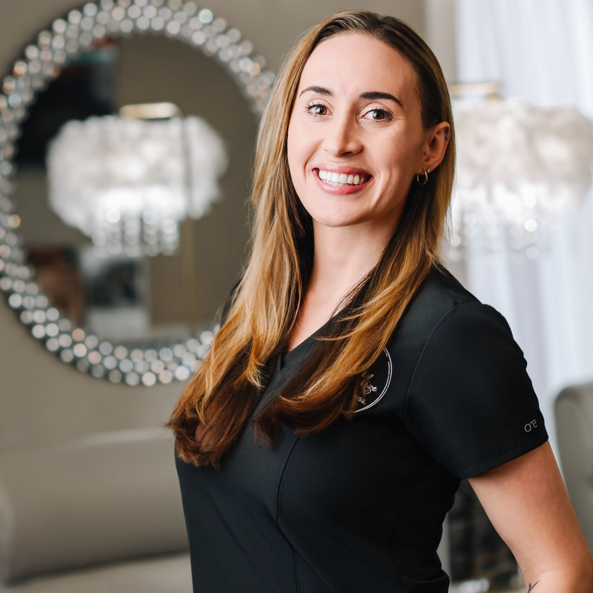 Smiling woman in a black top standing in a bright room with a round mirror and chandelier in the background