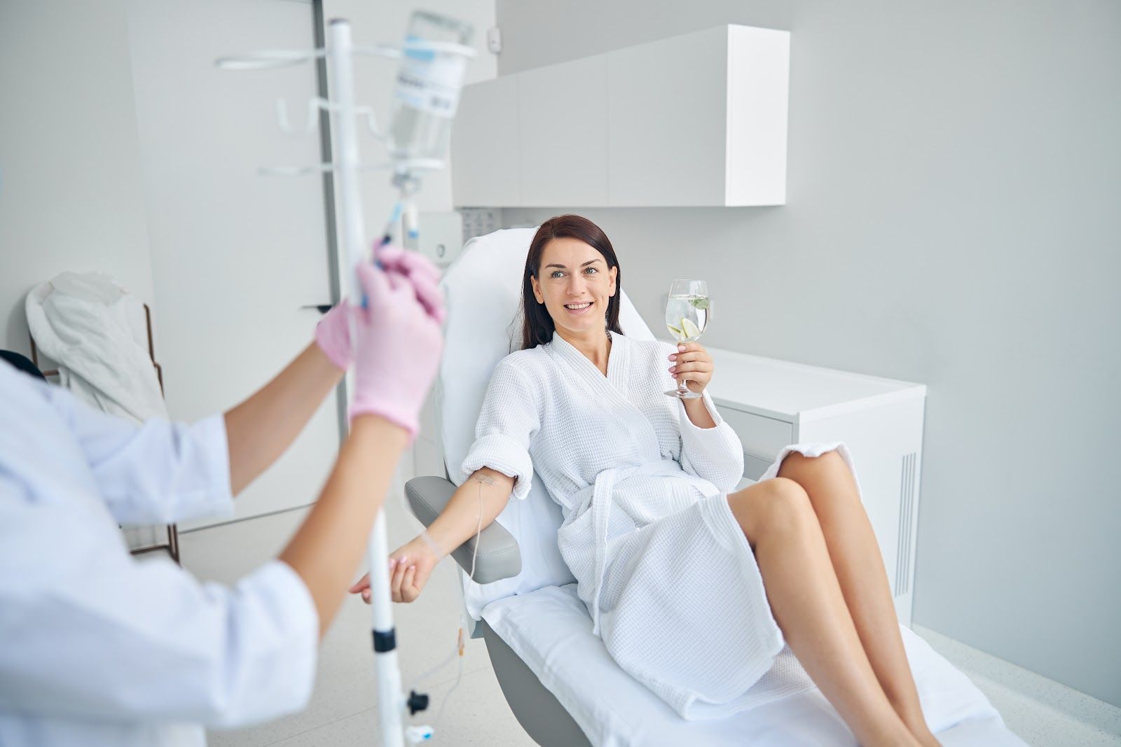 Woman in a spa chair holding a drink, with a staff member preparing an IV drip nearby.