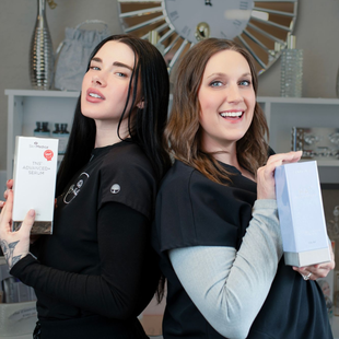 Two women stand back-to-back holding product boxes in front of a decorative wall clock.