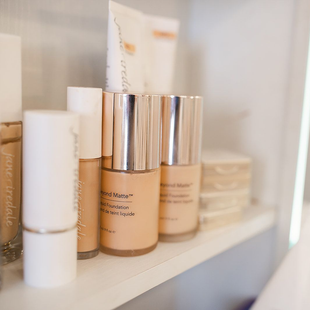 Assorted skincare bottles and jars on a white shelf, including beige and silver containers.