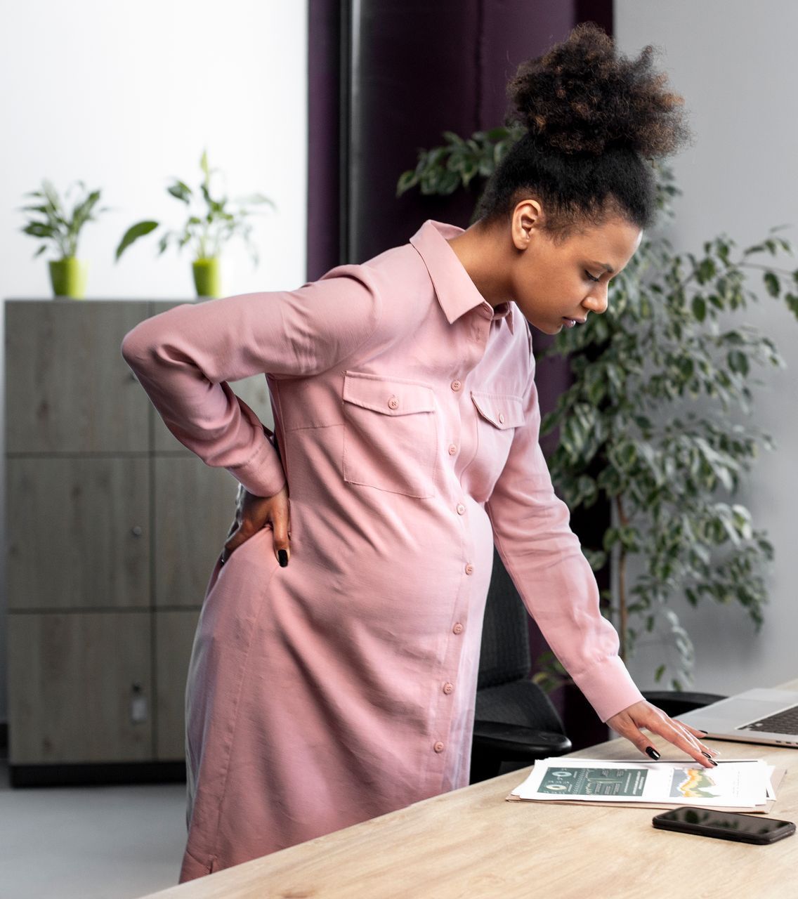 A pregnant woman is standing at a desk with her hands on her back.