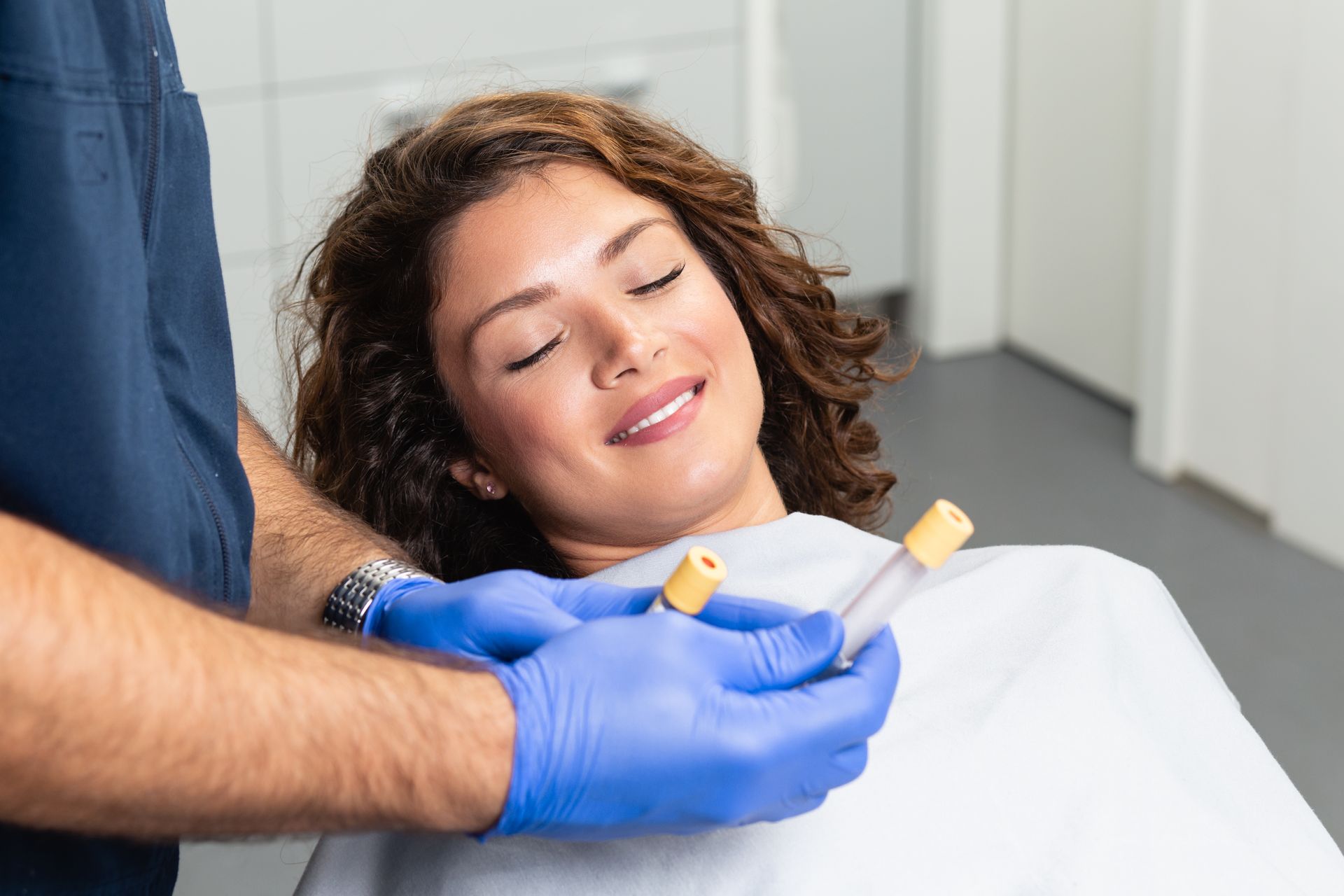 A woman is sitting in a dental chair with her eyes closed while a dentist holds two test tubes.