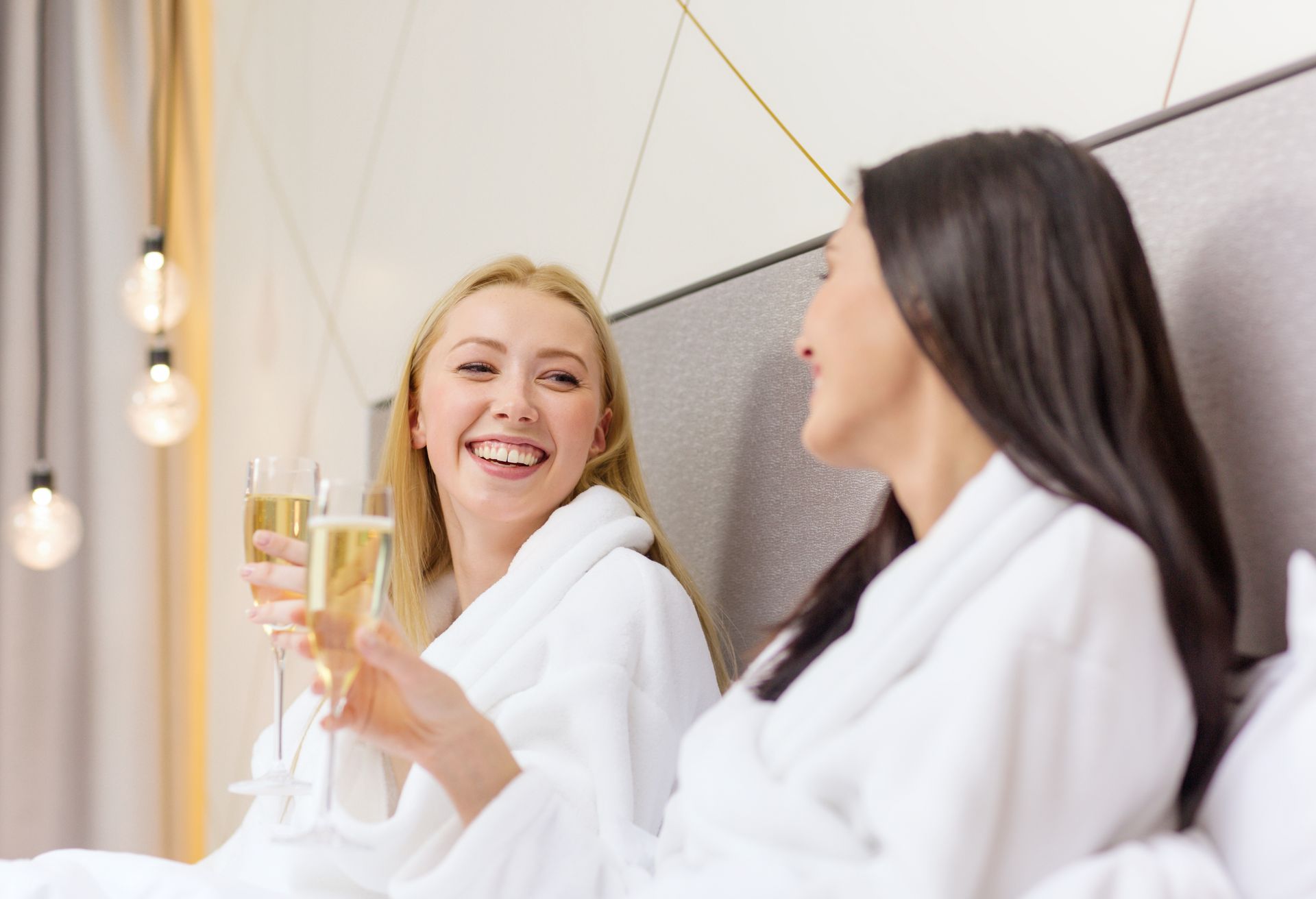 Two women in bathrobes are sitting on a bed drinking champagne.