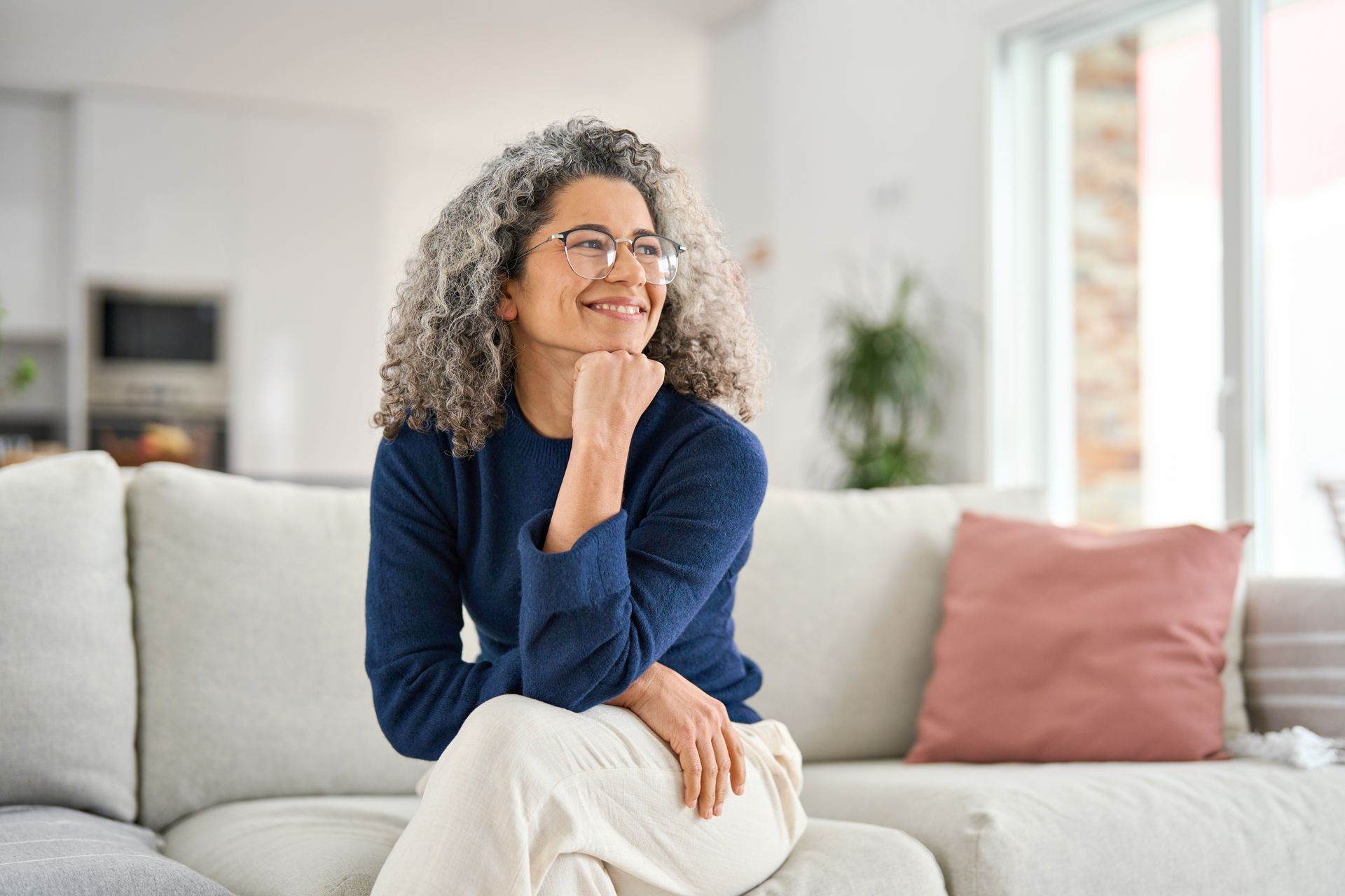 Smiling person sitting on a white sofa in a bright living room, wearing a navy top and light pants.