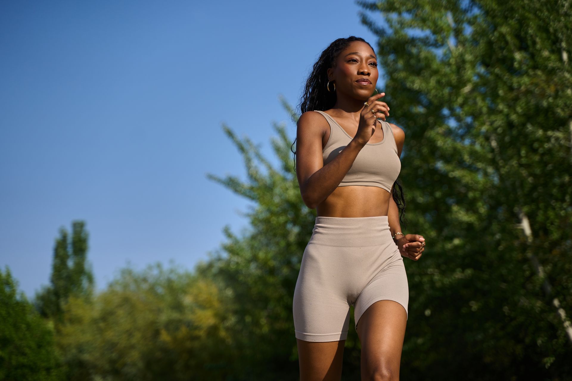Woman jogging in a beige workout outfit on a sunny outdoor path with trees and blue sky.
